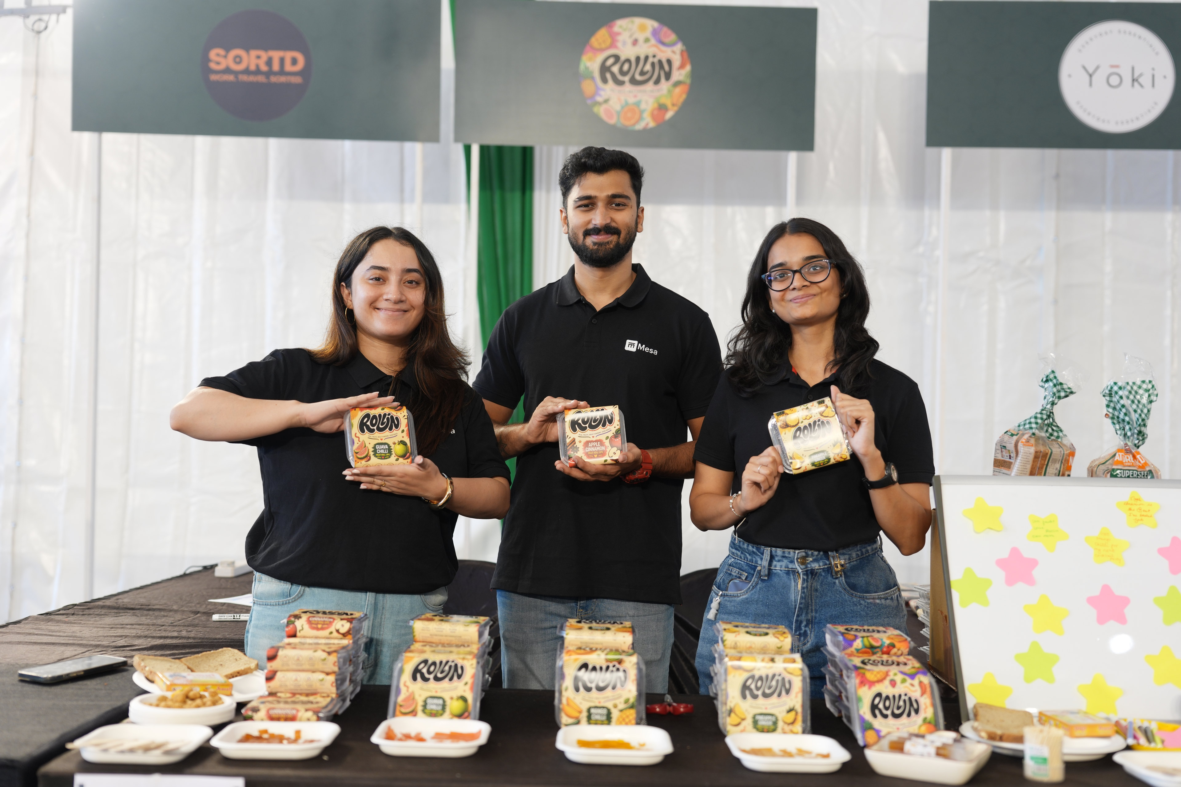 Three Mesa student founders holding up their Rollin packaged food products at a market stall