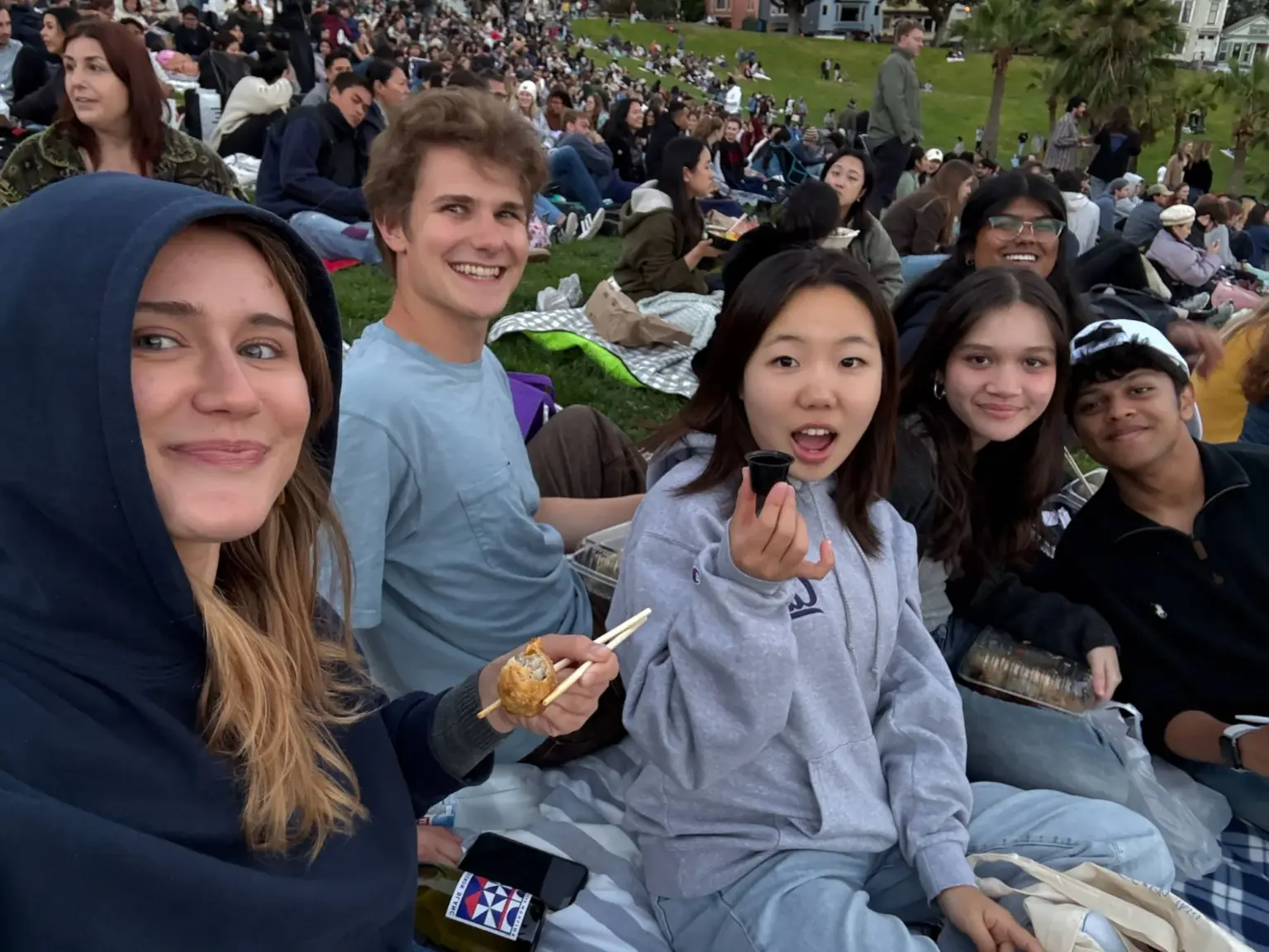 Mesa School undergraduate students sharing food on a picnic blanket in a crowded San Francisco park
